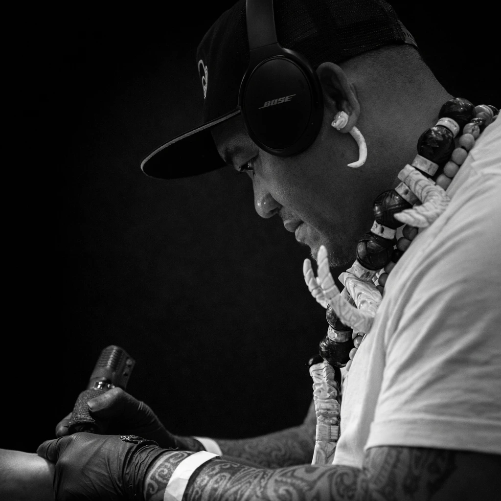 Portrait en noir et blanc de Nicolas Hikutini, tatoueur polynésien, concentré en train de tatouer, portant un collier marquisien en dents de cochon et un casque audio, éclairage dramatique sur fond sombre.